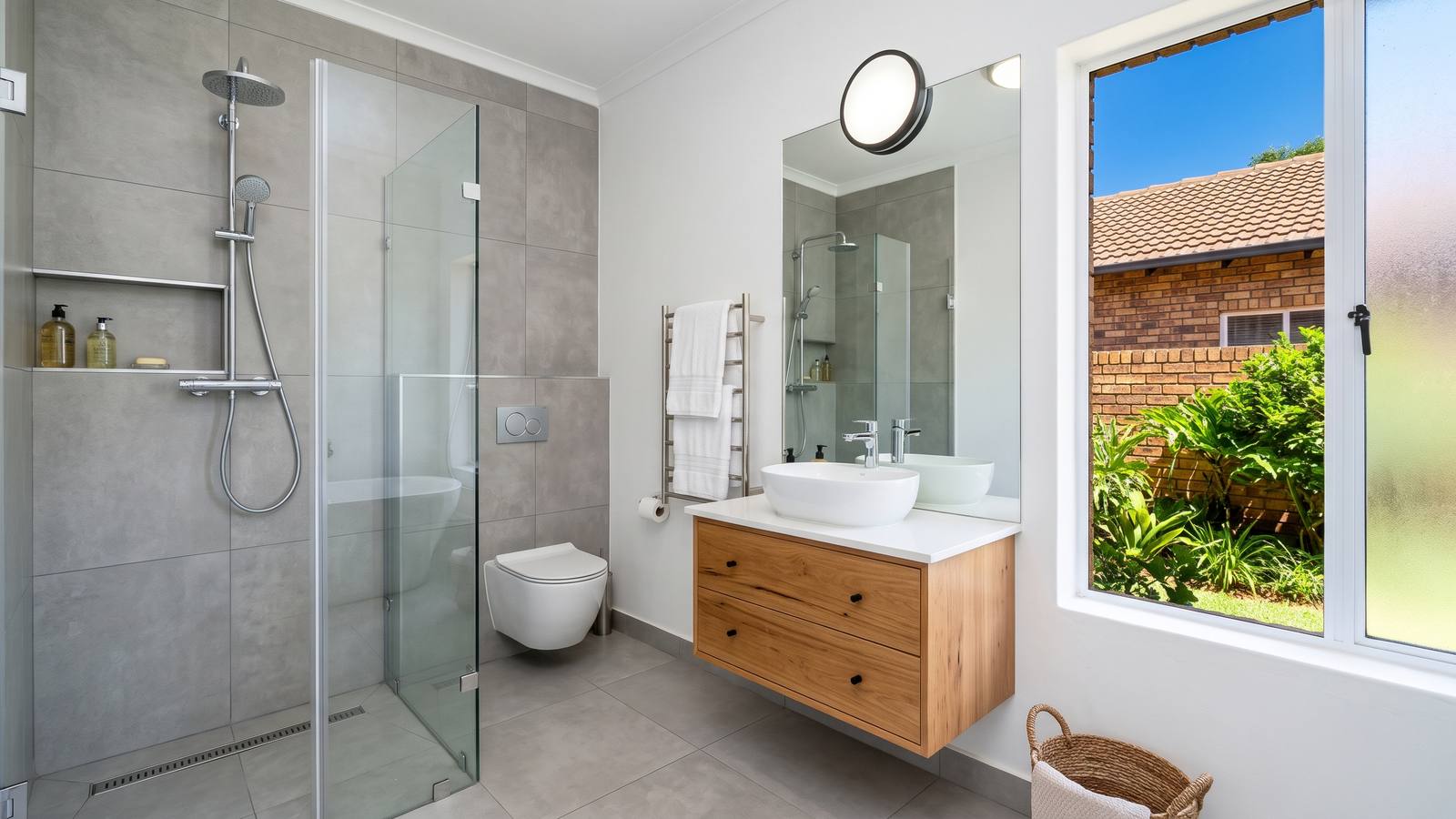 Bathroom with a frameless walk-in shower, grey wall tiles, a timber vanity and a wall-hung toilet.